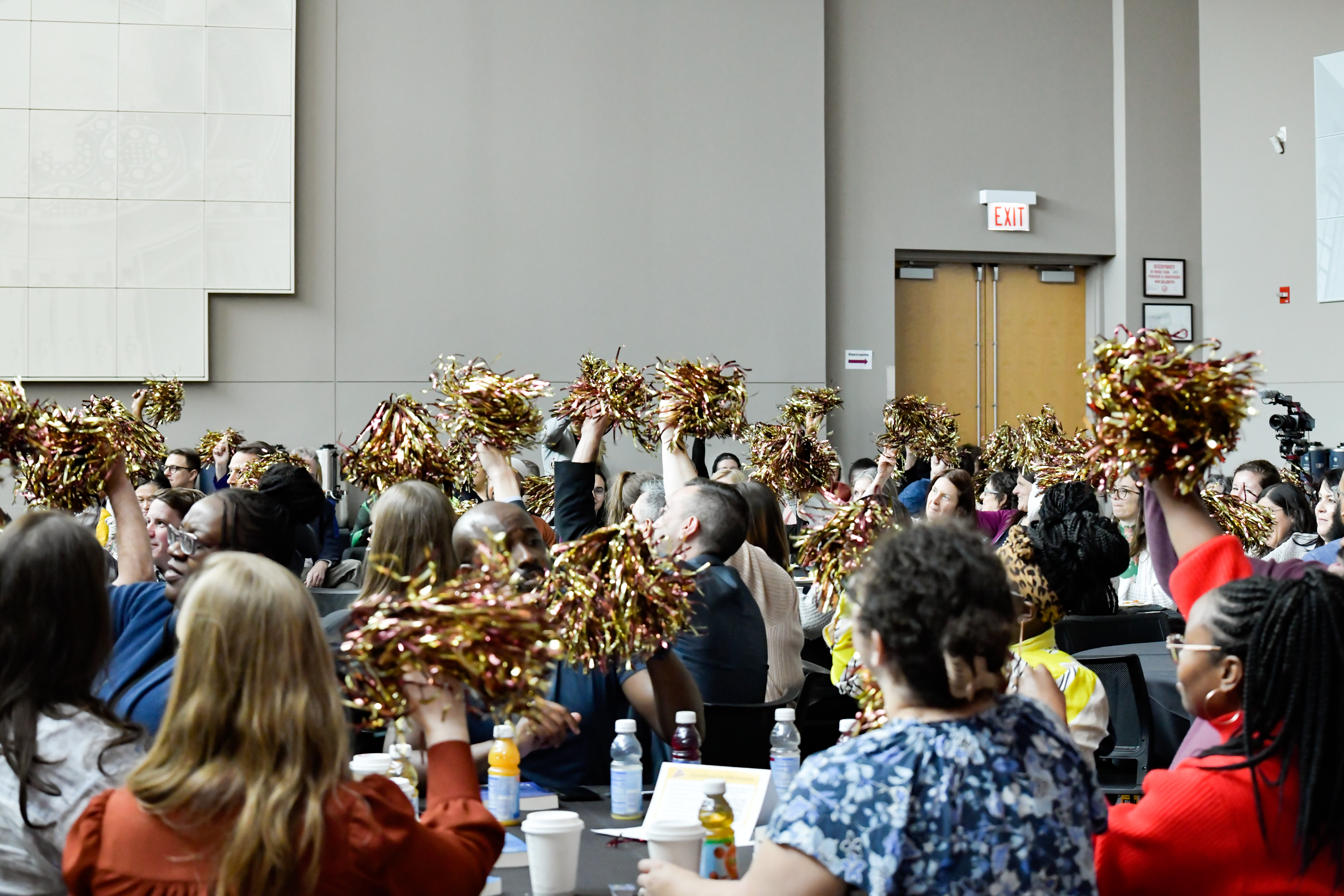 Group of faculty and staff sit at tables waving gold and maroon colored pom poms
